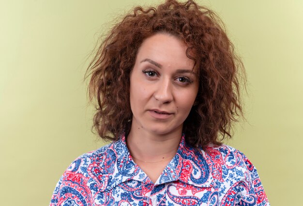 Young woman with short curly hair  in colorful shirt  looking with serious expression over green wall