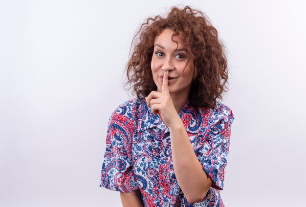 Young woman with short curly hair  in colorful shirt making silence gesture with fingers on lips smiling standing over white wall