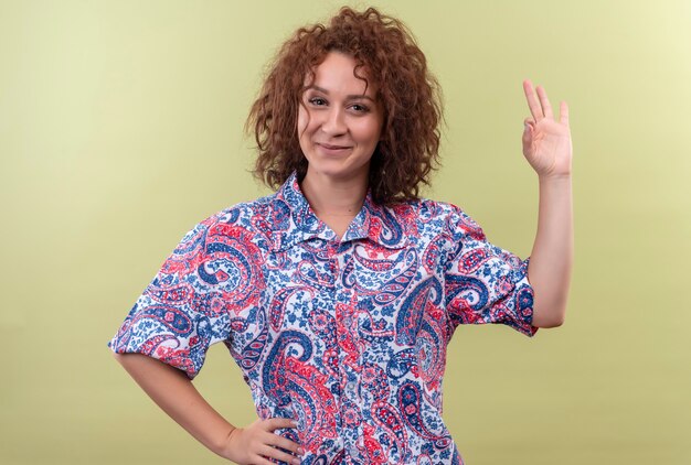 Young woman with short curly hair  in colorful shirt  smiling confident doing ok sign over green wall