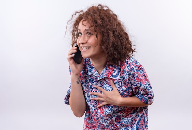 Молодая женщина с короткими вьющимися волосами в яркой рубашке разговаривает по мобильному телефону с застенчивой улыбкой на лице, стоя над белой стеной