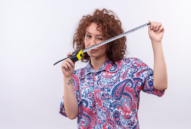 Young woman with short curly hair  in colorful shirt using measure tape looking suspicious standing over white wall