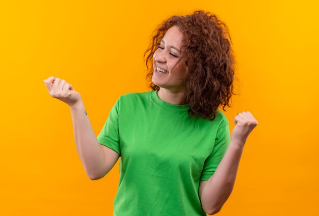 オレンジ色の壁の上に立って幸せで興奮した拳を握りしめている緑のTシャツの短い巻き毛の若い女性