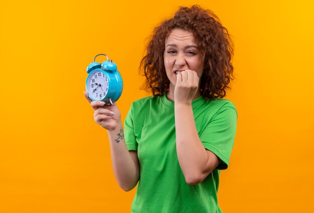 Young woman with short curly hair in green t-shirt holding alarm clock stressed and nervous biting nails standing over orange wall