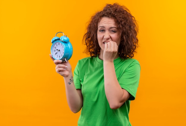 Young woman with short curly hair in green t-shirt holding alarm clock stressed and nervous biting nails standing over orange wall