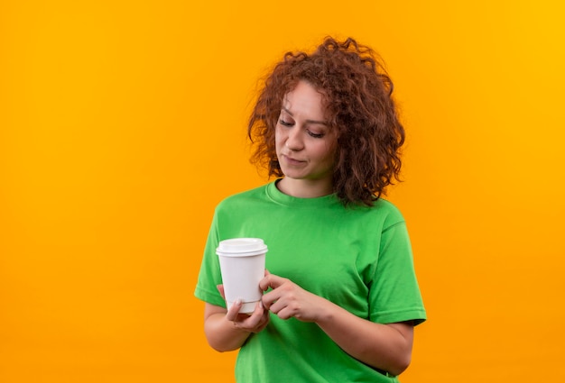 Young woman with short curly hair in green t-shirt holding coffee cup looking at it with sad expression on face standing over orange wall