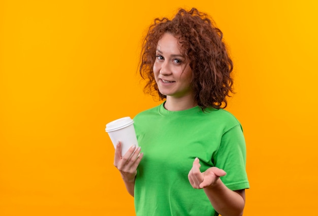 Giovane donna con i capelli ricci corti in maglietta verde che tiene la tazza di caffè che alza il braccio come fare domanda in piedi