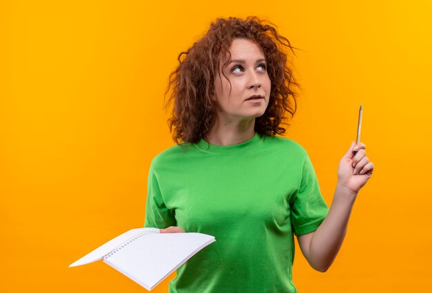 Young woman with short curly hair in green t-shirt holding notebook and pen looking up thinking standing