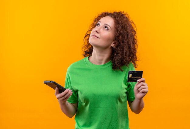 Young woman with short curly hair in green t-shirt holding two smartphones looking up with pensive expression on face trying to make choice standing