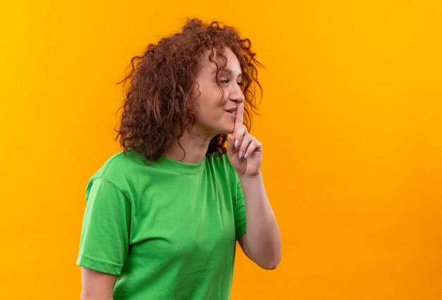 Young woman with short curly hair in green t-shirt looking aside making silence gesture with finger on lips standing