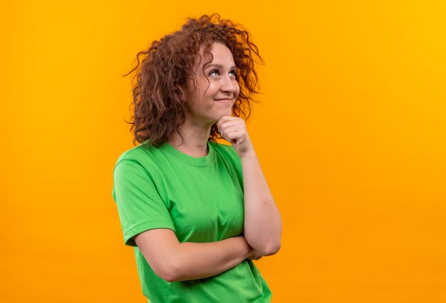 Молодая женщина с короткими вьющимися волосами в зеленой футболке смотрит в сторону с мечтательным взглядом и думает стоя