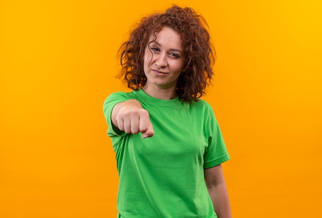 Young woman with short curly hair in green t-shirt looking confident pointing with index finger at front