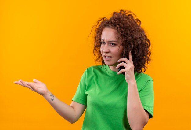 Молодая женщина с короткими вьющимися волосами в зеленой футболке выглядит смущенной и очень взволнованной во время разговора по мобильному телефону стоя