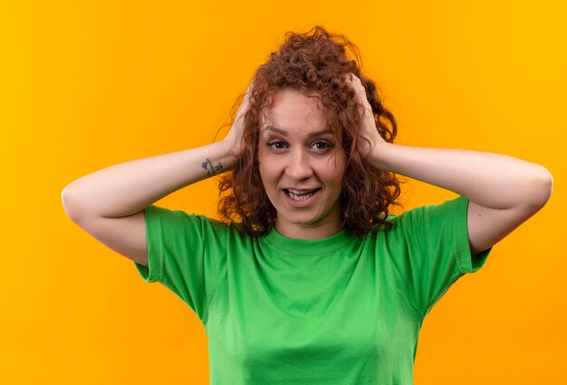 Young woman with short curly hair in green t-shirt looking surprised touching her hair smuling standing
