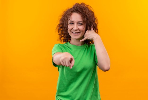 Young woman with short curly hair in green t-shirt making call me gesture pointing with finger at front