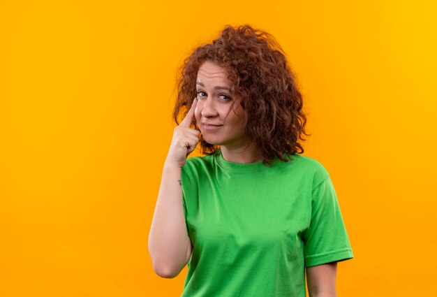 Young woman with short curly hair in green t-shirt pointing with index finger at eye watching you gesture standing over orange wall