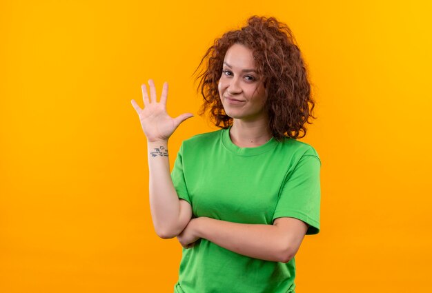 녹색 티셔츠에 짧은 곱슬 머리를 가진 젊은 여자가 오렌지 벽 위에 서있는 손으로 흔들며 웃고