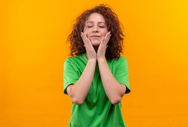 Молодая женщина с короткими вьющимися волосами в зеленой футболке, касаясь ее лица руками, испытывая положительные эмоции, стоя над оранжевой стеной