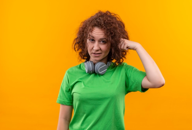 Молодая женщина с короткими вьющимися волосами в зеленой футболке с наушниками выглядит смущенной, указывая пальцем на висок, стоящий над оранжевой стеной