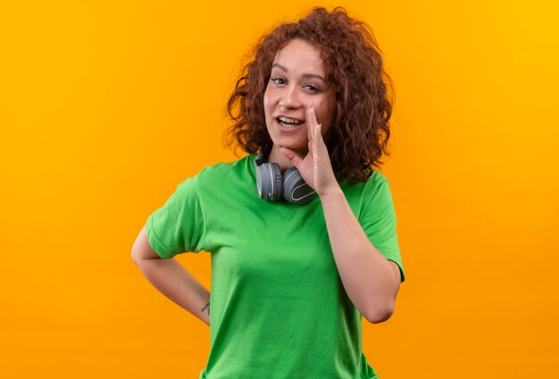 Young woman with short curly hair in green t-shirt with headphones telling a secret with hand near mouth standing over orange wall