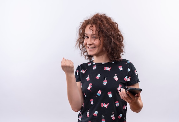 Young woman with short curly hair holding smartphone clenching fist happy and excited rejoicing her success standing over white wall