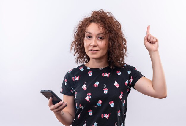 Young woman with short curly hair holding smartphone pointing up with index finger looking smart having great idea standing over white wall