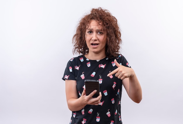 Young woman with short curly hair holding smartphone pointing with finger to it emotional and stressed standing over white wall