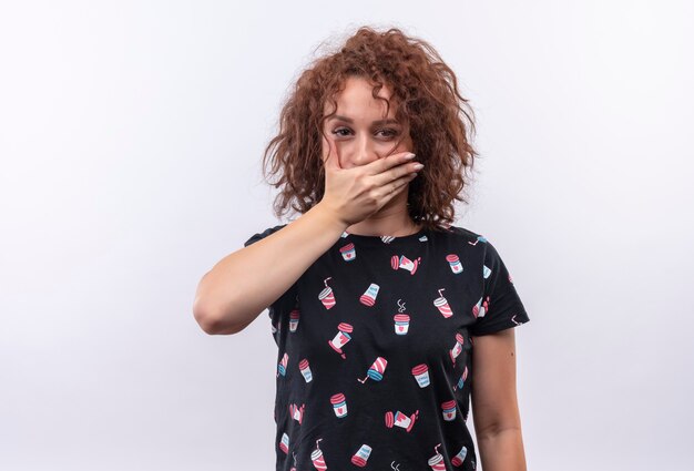 Foto gratuita giovane donna con capelli ricci corti guardando stupito che copre la bocca con la mano in piedi sul muro bianco