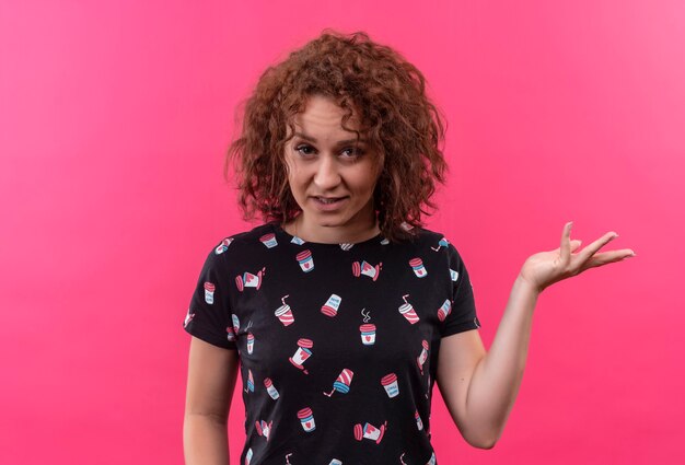 Young woman with short curly hair looking arguing with raised arm standing over pink wall