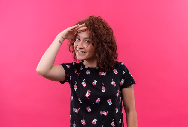 Free photo young woman with short curly hair looking far away with hand over head to look someone or something standing over pink wall