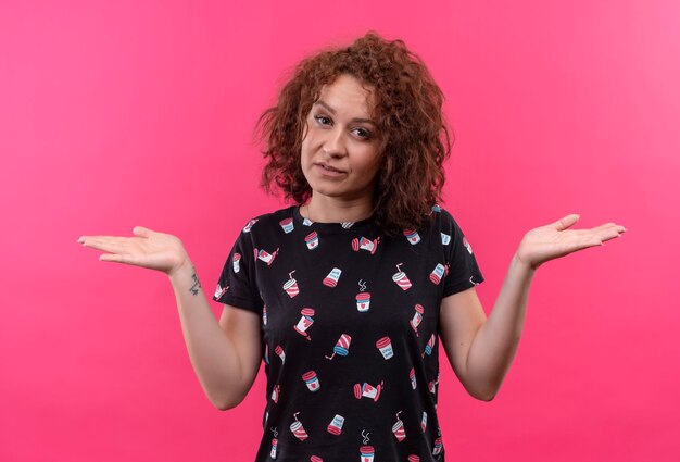 Young woman with short curly hair looking uncertain and confused shrugging shoulders, having no answer standing over pink wall