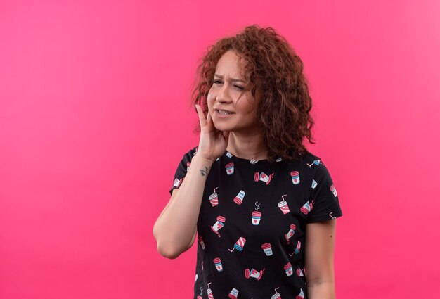 Young woman with short curly hair looking unwell touching cheek having toothache standing over pink wall