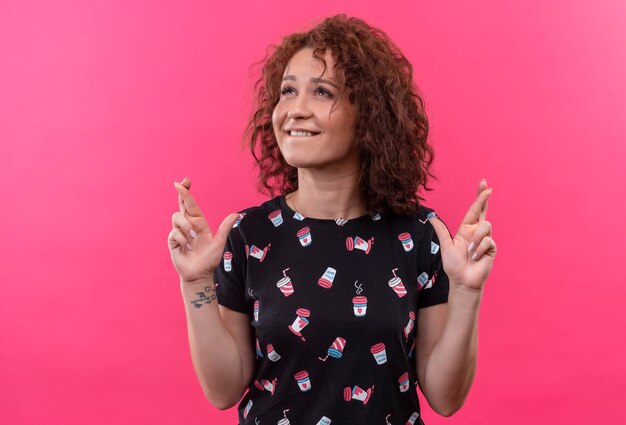 Young woman with short curly hair making desirable wish crossing fingers with hope expression standing over pink wall