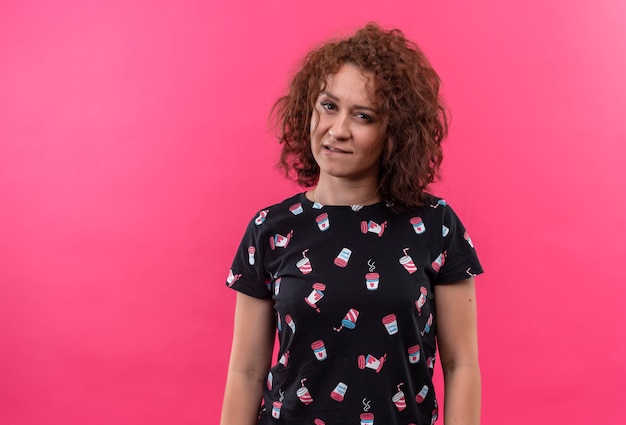 Young woman with short curly hair smiling looking biting lip seductively standing over pink wall