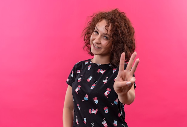 Бесплатное фото Молодая женщина с короткими вьющимися волосами улыбается, показывая пальцами номер три и указывая вверх, стоя над розовой стеной