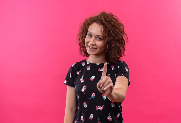 Young woman with short curly hair smiling showing index finger warning standing over pink wall