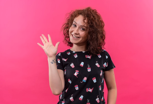 Free photo young woman with short curly hair smiling waving with hand standing over pink wall