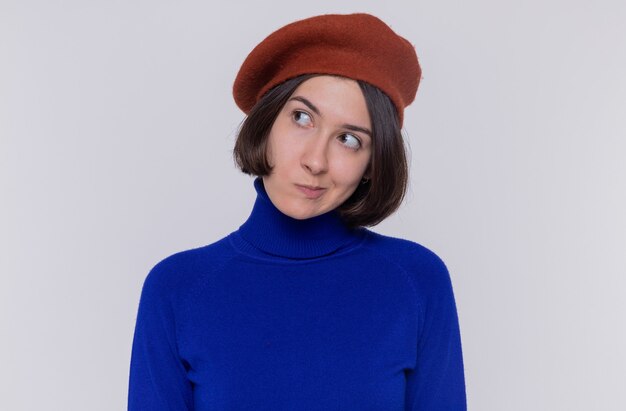 Young woman with short hair in blue turtleneck wearing beret looking aside with skeptic expression standing over white wall