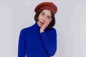 Free photo young woman with short hair in blue turtleneck wearing beret looking at front confused and surprised standing over white wall