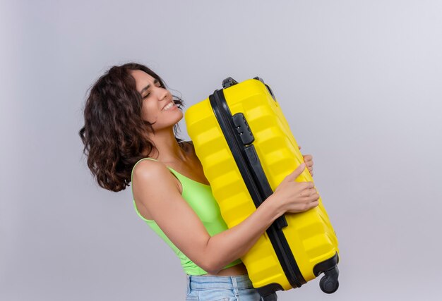 A young woman with short hair in green crop top carrying heavy yellow suitcase on a white background