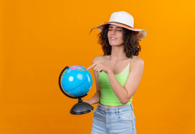 A young woman with short hair in green crop top wearing sun hat pointing at a globe with index finger 