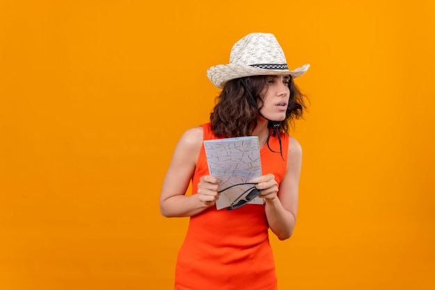 オレンジ色のシャツを着た短い髪の若い女性は、サングラスをかけた地図を保持している太陽の帽子をかぶっています