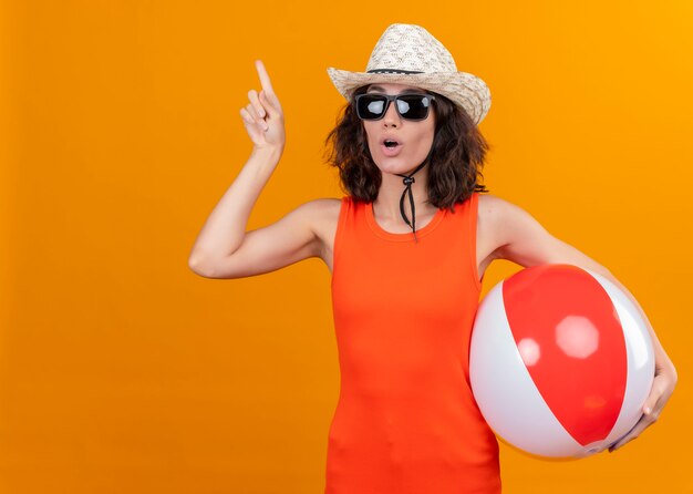 A young woman with short hair in an orange shirt wearing sun hat and sunglasses holding inflatable ball pointing at right side with index finger 