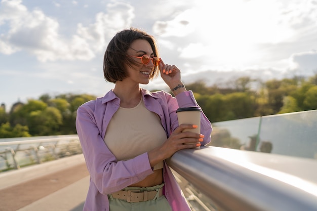 スタイリッシュな夏の衣装で短い髪の若い女性は、現代の橋でコーヒーを飲みます
