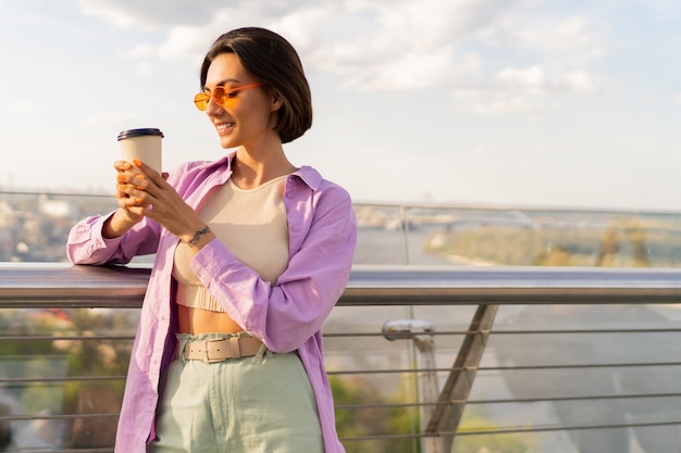 スタイリッシュな夏の衣装で短い髪の若い女性は、現代の橋でコーヒーを飲みます