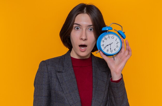 Young woman with short hair wearing grey jacket holding alarm clock looking at front amazed and surprised standing over orange wall