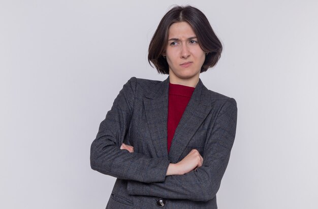 Free photo young woman with short hair wearing grey jacket looking aside with skeptic expression with arms crossed standing over white wall