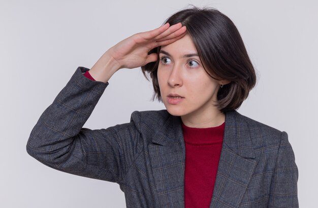 Young woman with short hair wearing grey jacket looking far away with hand over head to look someone or something