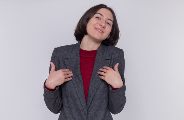 Young woman with short hair wearing grey jacket looking at front smiling confident pointing at herself standing over white wall