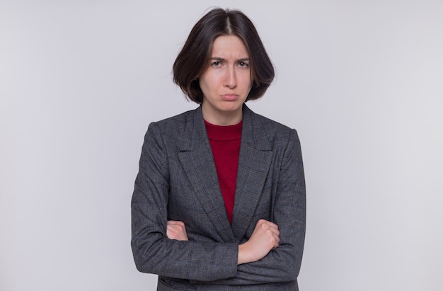 Young woman with short hair wearing grey jacket looking at front with frowning face being offended with arms crossed standing over white wall