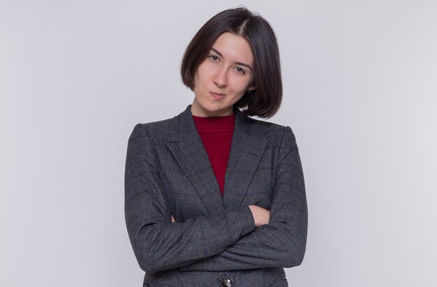 Young woman with short hair wearing grey jacket looking at front with skeptic expression standing over white wall